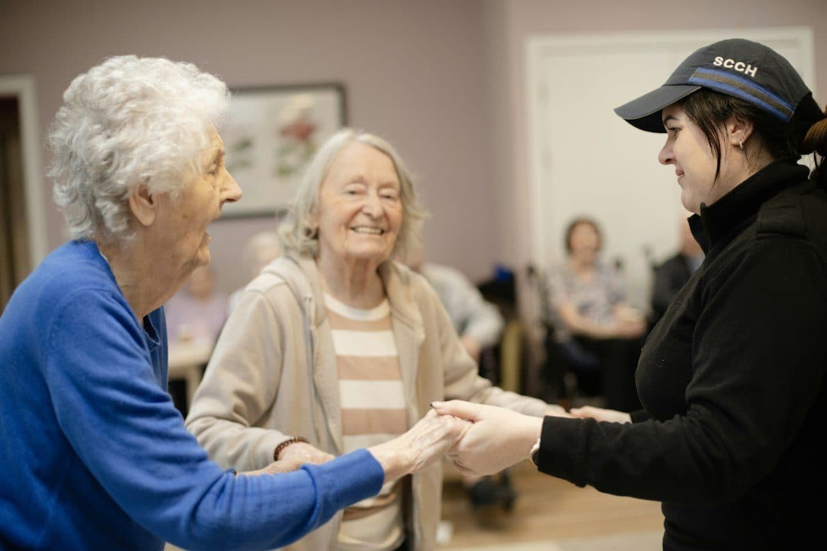 Two older women holding hands with a younger caregiver in a community room